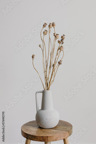 A ceramic vase on a wooden stool with dried poppy heads on branch. Minimal home decoration, copy space. White background, simple decor. 