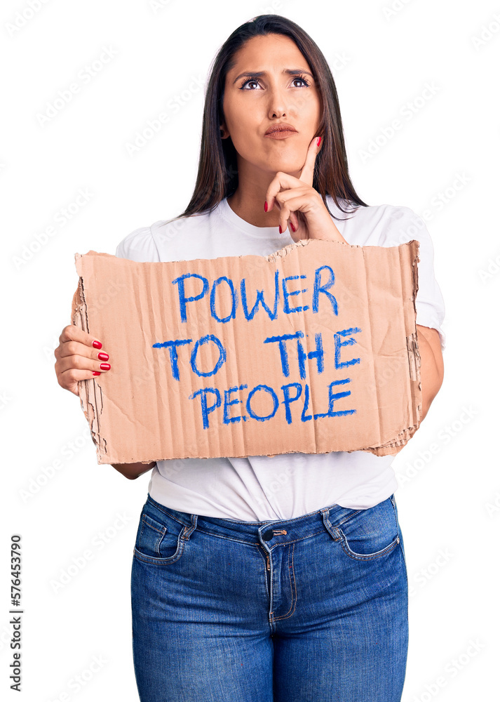 Young beautiful woman holding power to the people cardboard banner serious face thinking about question with hand on chin, thoughtful about confusing idea Young beautiful woman holding power to the people cardboard banner serious face thinking about question with hand on chin, thoughtful about confusing idea