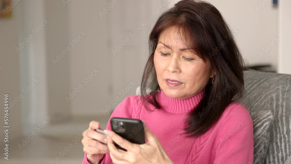 Smiling attractive woman in pink enjoying shopping online using mobile phone excited and happy, waving e-commerce credit card at success of placing internet order sitting in living room chair. Smiling attractive woman in pink enjoying shopping online using mobile phone excited and happy, waving e-commerce credit card at success of placing internet order sitting in living room chair.