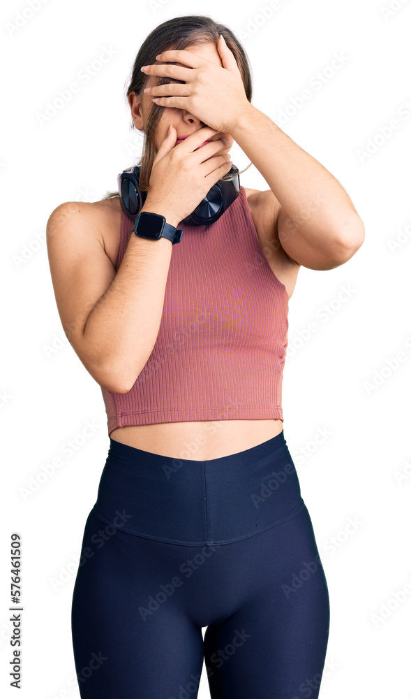 Beautiful caucasian young woman wearing gym clothes and using headphones covering eyes and mouth with hands, surprised and shocked. hiding emotion Beautiful caucasian young woman wearing gym clothes and using headphones covering eyes and mouth with hands, surprised and shocked. hiding emotion