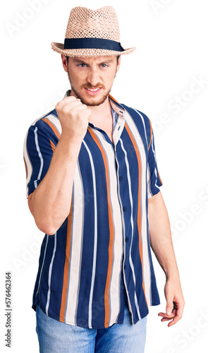 Young handsome caucasian man wearing summer hat angry and mad raising fist frustrated and furious while shouting with anger. rage and aggressive concept.