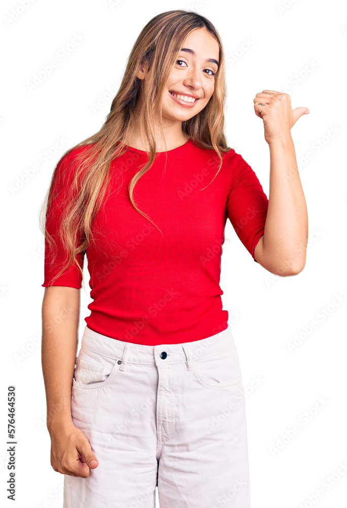 Young beautiful blonde woman wearing casual clothes smiling with happy face looking and pointing to the side with thumb up. Young beautiful blonde woman wearing casual clothes smiling with happy face looking and pointing to the side with thumb up.
