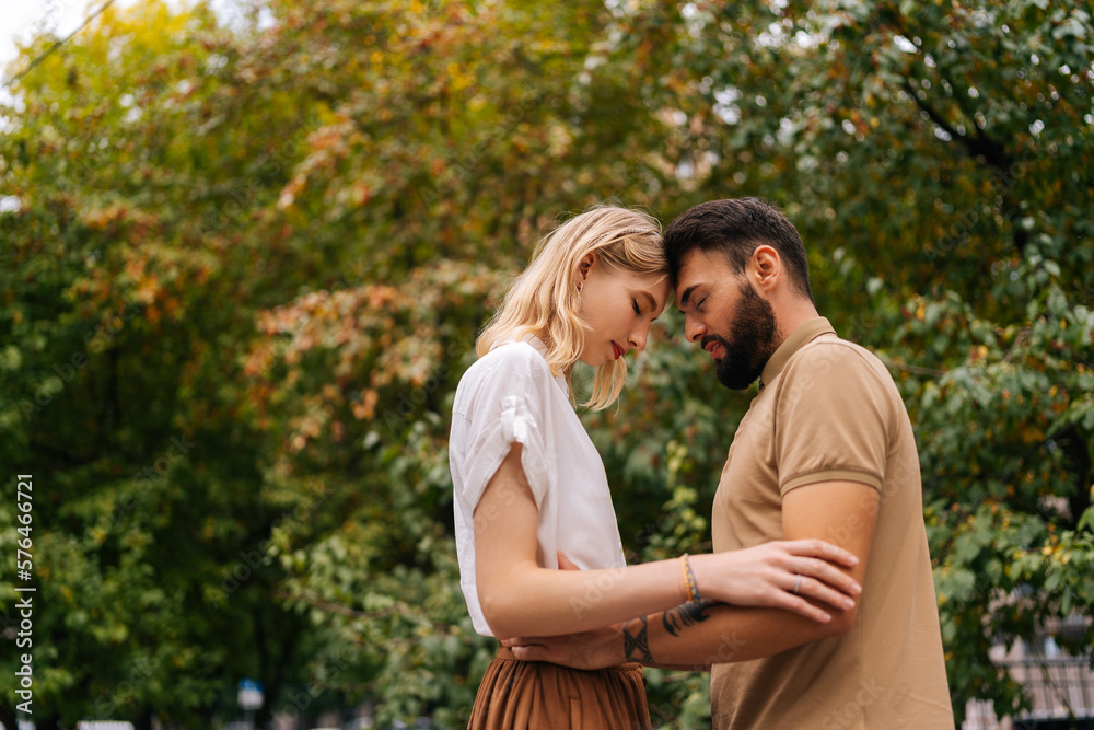 Low-angle view of happy young couple in love standing with closed eyes in front of each other at city park on background of trees. Tattooed handsome man and pretty lady walking spending time together. Low-angle view of happy young couple in love standing with closed eyes in front of each other at city park on background of trees. Tattooed handsome man and pretty lady walking spending time together.