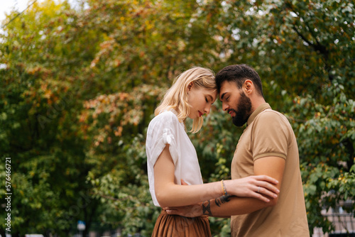 Low-angle view of happy young couple in love standing with closed eyes in front of each other at city park on background of trees. Tattooed handsome man and pretty lady walking spending time together.