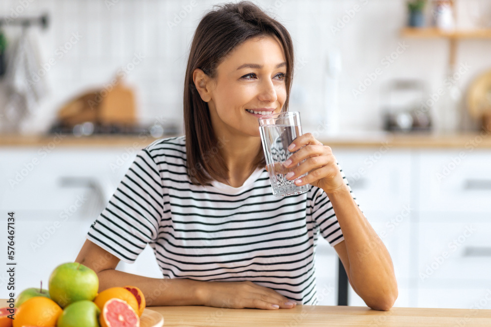 Portrait of a beautiful woman drinking water sitting at a table in the kitchen looking away and smiling, wellness healthy food concept Portrait of a beautiful woman drinking water sitting at a table in the kitchen looking away and smiling, wellness healthy food concept