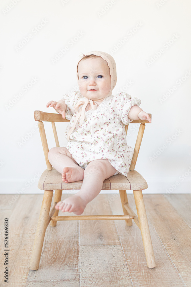 baby with a limb difference disability sitting in a wooden chair with a bonnet and floral dress baby with a limb difference disability sitting in a wooden chair with a bonnet and floral dress