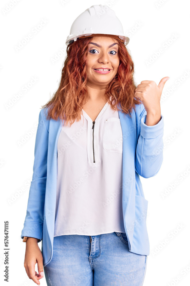 Young latin woman wearing architect hardhat smiling with happy face looking and pointing to the side with thumb up. Young latin woman wearing architect hardhat smiling with happy face looking and pointing to the side with thumb up.