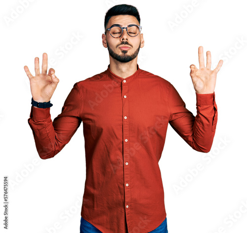Young hispanic man with beard wearing business shirt and glasses relax and smiling with eyes closed doing meditation gesture with fingers. yoga concept. © Krakenimages.com