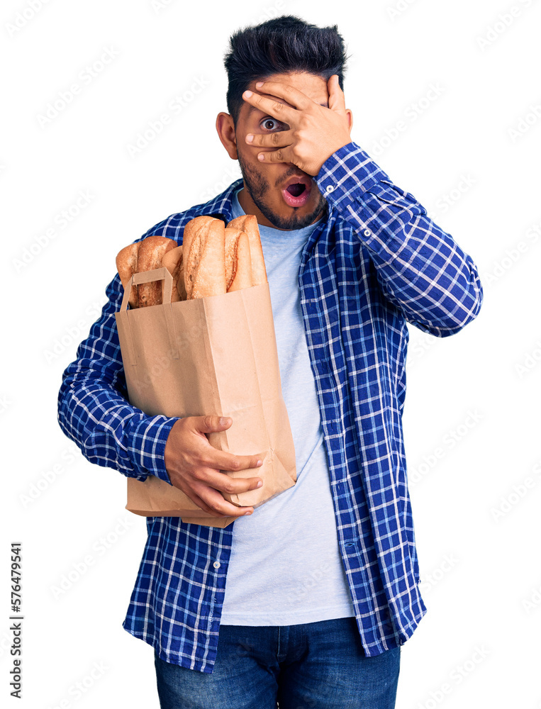Handsome latin american young man holding paper bag with bread peeking in shock covering face and eyes with hand, looking through fingers with embarrassed expression. Handsome latin american young man holding paper bag with bread peeking in shock covering face and eyes with hand, looking through fingers with embarrassed expression.
