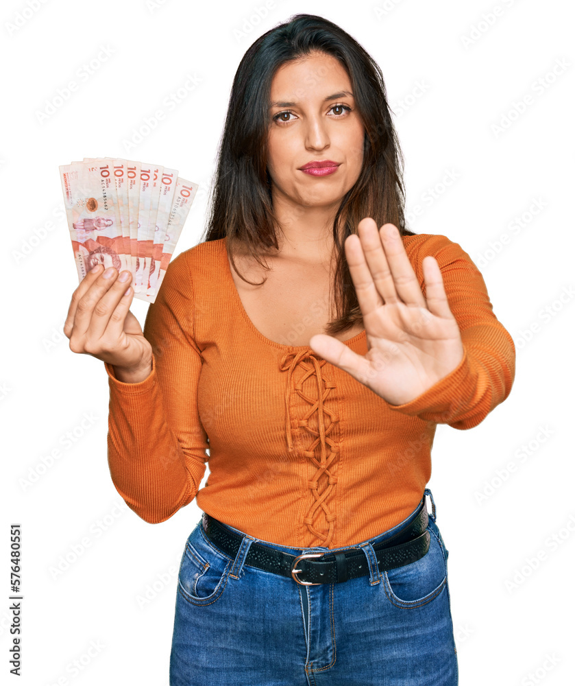 Beautiful hispanic woman holding 10 colombian pesos banknotes with open hand doing stop sign with serious and confident expression, defense gesture Beautiful hispanic woman holding 10 colombian pesos banknotes with open hand doing stop sign with serious and confident expression, defense gesture
