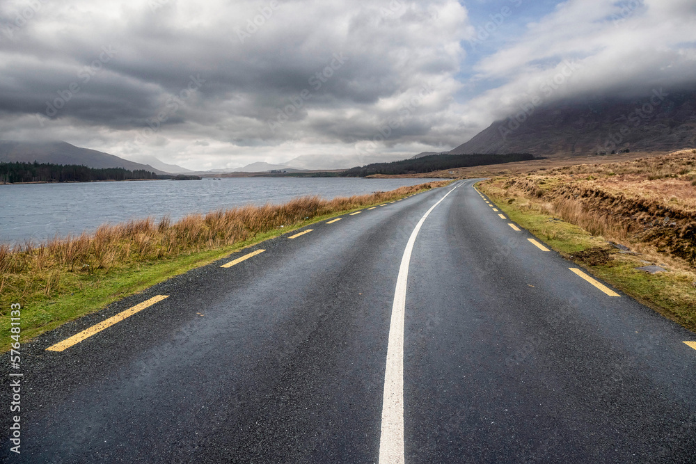 Beautiful scenery and small road by a lake in Connemara, Ireland. Mountains and cloudy sky in the background. Travel and transportation concept.