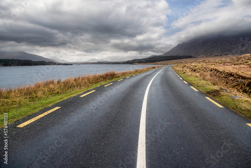 Beautiful scenery and small road by a lake in Connemara, Ireland. Mountains and cloudy sky in the background. Travel and transportation concept.