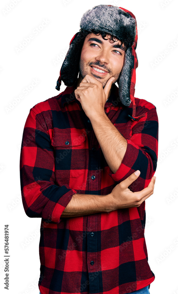 Hispanic young man with beard wearing fluffy earmuff hat with hand on chin thinking about question, pensive expression. smiling with thoughtful face. doubt concept. Hispanic young man with beard wearing fluffy earmuff hat with hand on chin thinking about question, pensive expression. smiling with thoughtful face. doubt concept.
