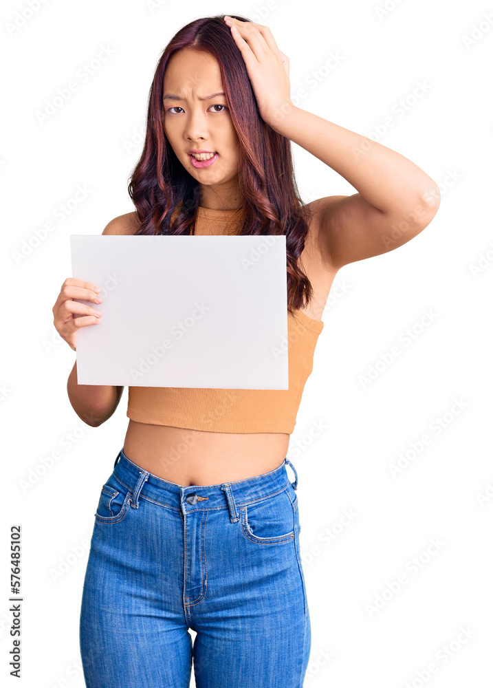 Young beautiful chinese girl holding paper banner with blank space stressed and frustrated with hand on head, surprised and angry face Young beautiful chinese girl holding paper banner with blank space stressed and frustrated with hand on head, surprised and angry face