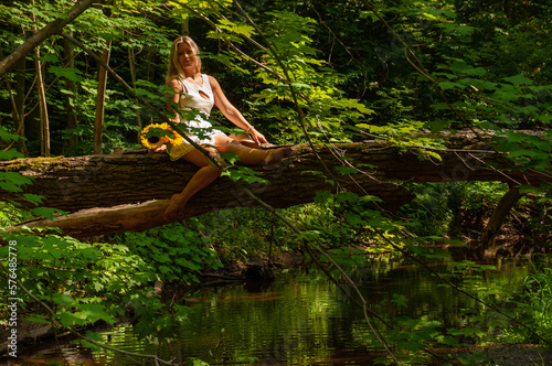 A woman in a white dress holds sunflowers as she sits on a fallen tree over a creek