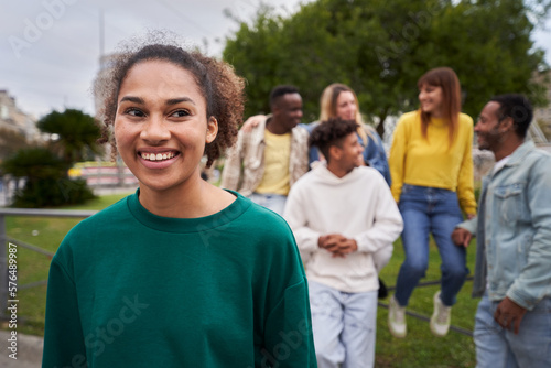 Young Latin girl with wide smile and looking away. Group of friends gathered in the background of the image. Colleagues hanging out in the city outdoors.