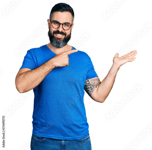 Hispanic man with beard wearing casual t shirt and glasses amazed and smiling to the camera while presenting with hand and pointing with finger.