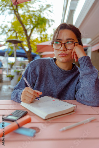 joven mujer, de 20 años, sentada en un cafe, con un libro, anotando ideas y estudiando enojada.
