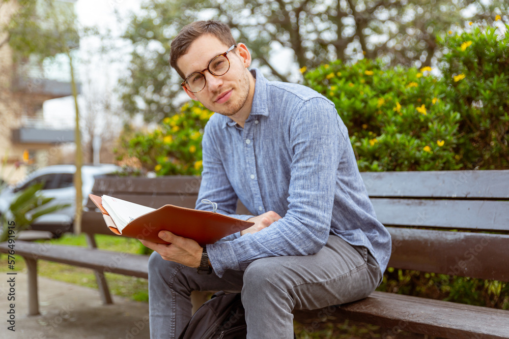 Obraz premium Hombre joven, caucásico, de 25 años, leyendo un libro, en un banco de un parque.