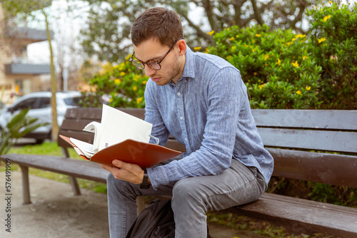 Hombre joven, caucásico, de 25 años, leyendo un libro, en un banco de un parque.