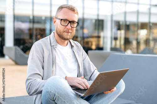 Positive attractive clever caucasian man with a glasses, in a casual clothes, IT specialist, developer, programmer, sit with a laptop outdoors, developing a new program, writing code, smiles at camera
