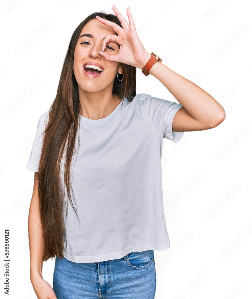 Young hispanic girl wearing casual white t shirt doing ok gesture with hand smiling, eye looking through fingers with happy face. Young hispanic girl wearing casual white t shirt doing ok gesture with hand smiling, eye looking through fingers with happy face.