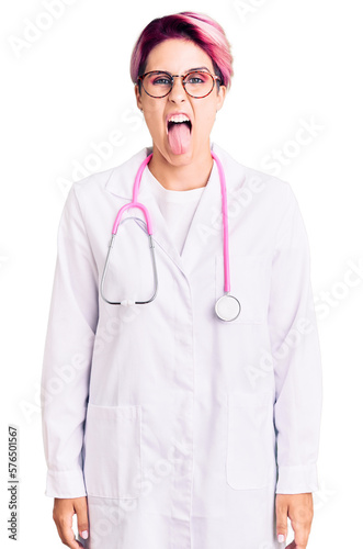 Young beautiful woman with pink hair wearing doctor uniform sticking tongue out happy with funny expression. emotion concept.