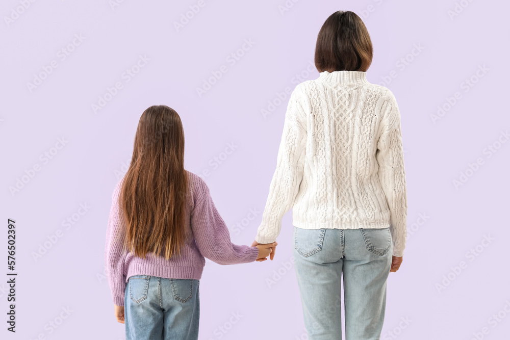 Little girl and her mother in warm sweaters holding hands on lilac background, back view Little girl and her mother in warm sweaters holding hands on lilac background, back view