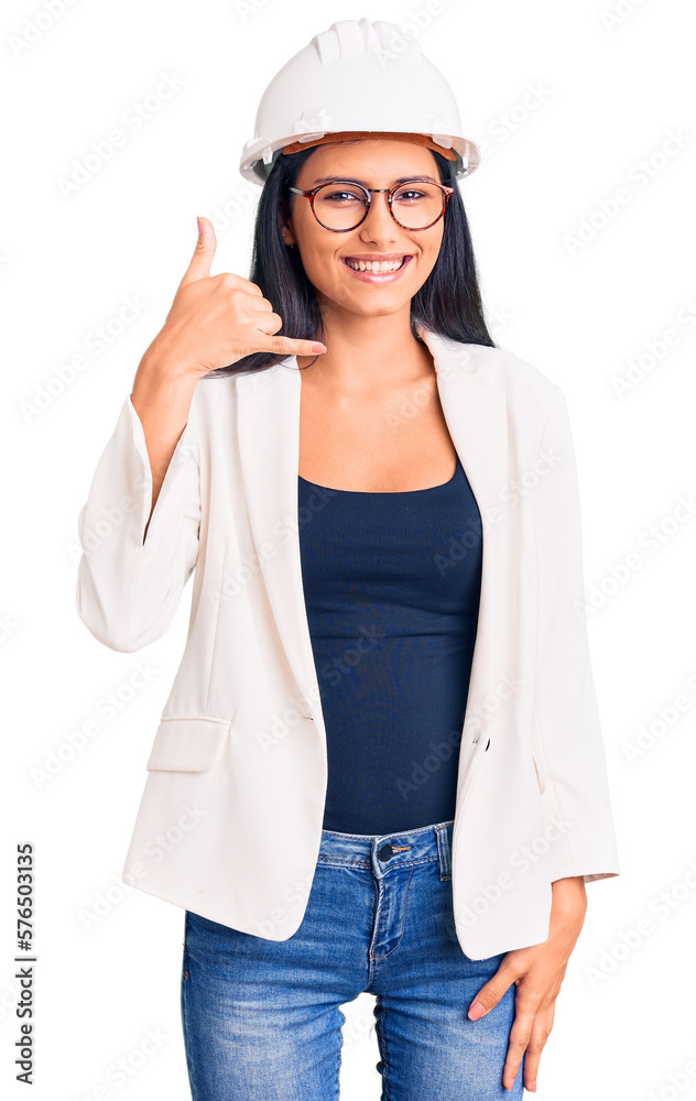 Young beautiful latin girl wearing architect hardhat and glasses smiling doing phone gesture with hand and fingers like talking on the telephone. communicating concepts. Young beautiful latin girl wearing architect hardhat and glasses smiling doing phone gesture with hand and fingers like talking on the telephone. communicating concepts.