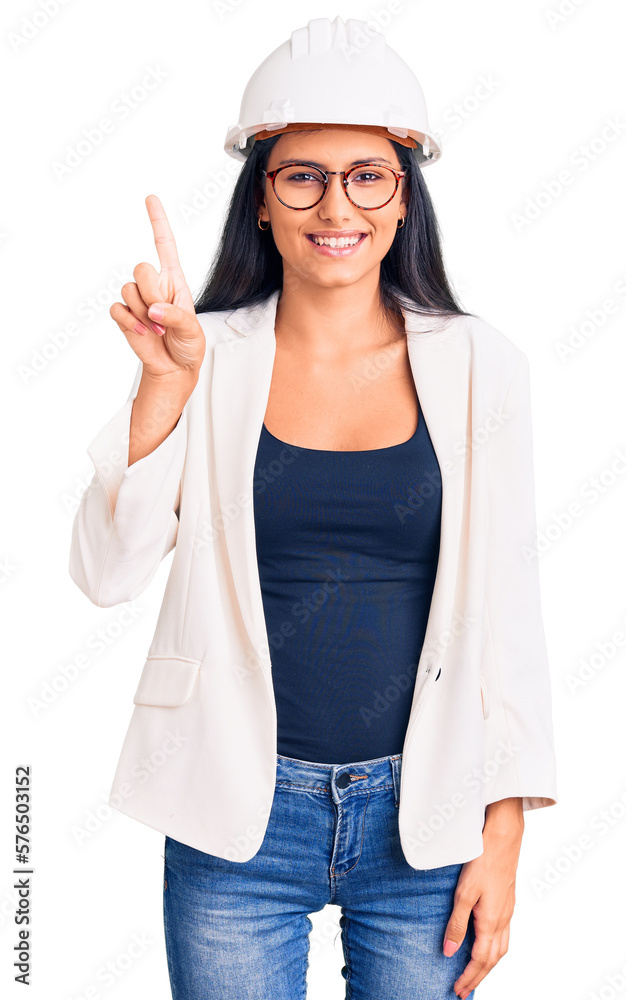 Young beautiful latin girl wearing architect hardhat and glasses showing and pointing up with finger number one while smiling confident and happy. Young beautiful latin girl wearing architect hardhat and glasses showing and pointing up with finger number one while smiling confident and happy.