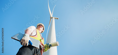 Low angle view of Asian engineer or architect working with his laptop in hand while supervising construction site in Wind turbine farm power generator in beautiful nature landscape. copy space.