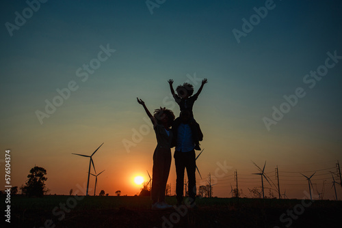 Silhouettes of Happy family father, mother and child daughter sits on the shoulders of his father with Wind turbine farm power generator in beautiful nature landscape for production of renewable green