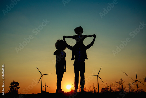 Silhouettes of Happy family father  mother and child daughter sits on the shoulders of his father with windmills for electricity generation at sunrise by producing sustainable energy 