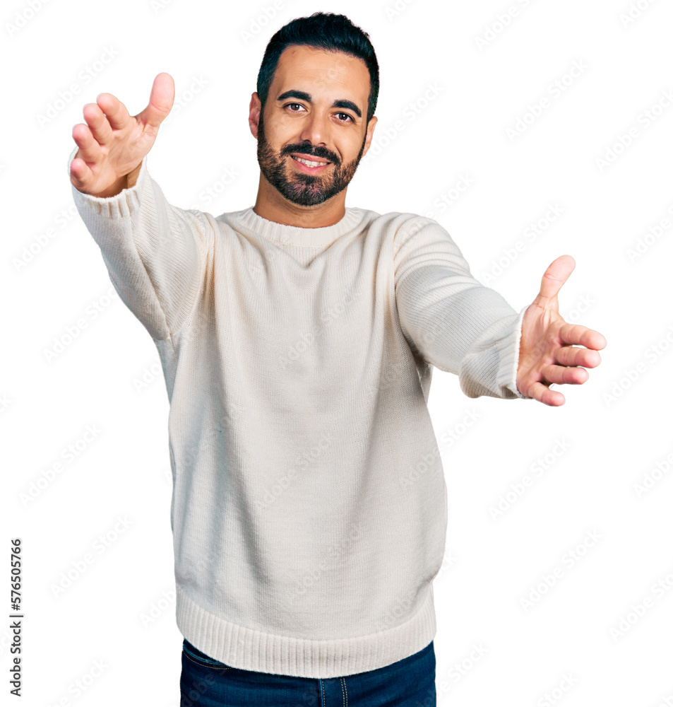 Naklejka premium Young hispanic man with beard wearing casual white sweater looking at the camera smiling with open arms for hug. cheerful expression embracing happiness.