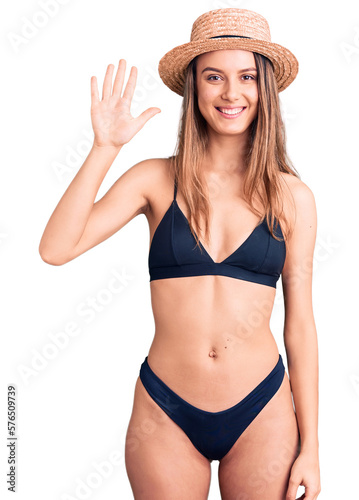 Young beautiful girl wearing bikini and hat showing and pointing up with fingers number five while smiling confident and happy.