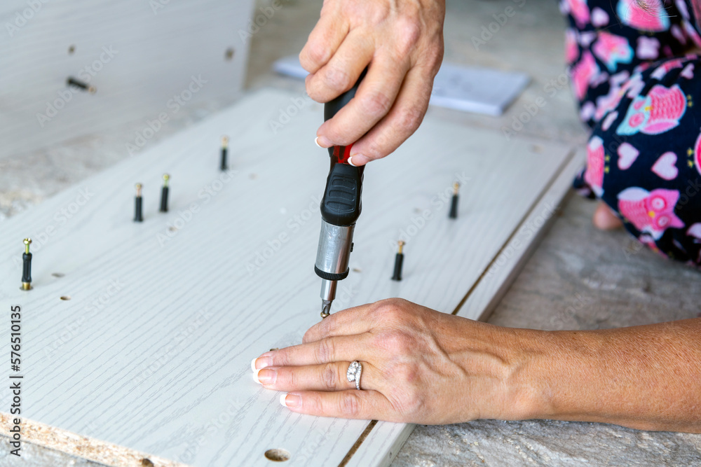 A woman sits on a rug and uses a ratchet screwdriver as she screws cam bolts, or camber adjustment bolts into pre-fabricated wood while assembling a piece of furniture. A woman sits on a rug and uses a ratchet screwdriver as she screws cam bolts, or camber adjustment bolts into pre-fabricated wood while assembling a piece of furniture.