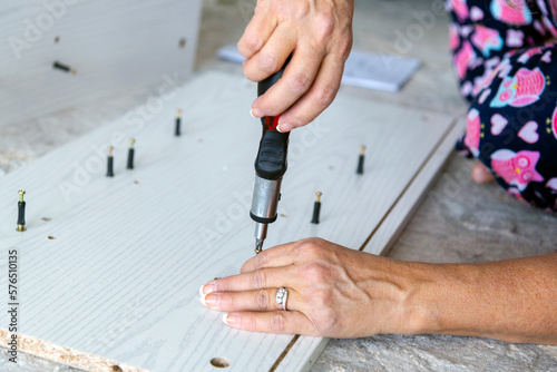 A woman sits on a rug and uses a ratchet screwdriver as she screws cam bolts, or camber adjustment bolts into pre-fabricated wood while assembling a piece of furniture.