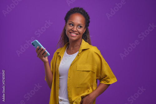 Young cheerful African American woman dressed in casual style using smartphone and looking around smiling enjoying watching social media feed posing on isolated lilac background. Phone, apps, Internet