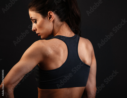 Serious female sporty muscular with ponytail doing stretching workout the shoulders, blades and arms and looking down in sport bra, standing on dark shadow grey background with empty space.