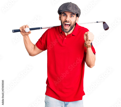 Young handsome man with beard playing golf holding club and ball screaming proud, celebrating victory and success very excited with raised arms