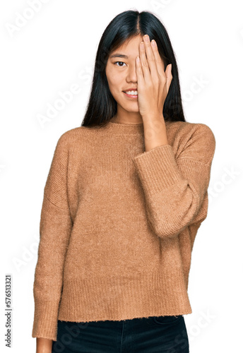 Beautiful young asian woman wearing casual winter sweater covering one eye with hand, confident smile on face and surprise emotion.