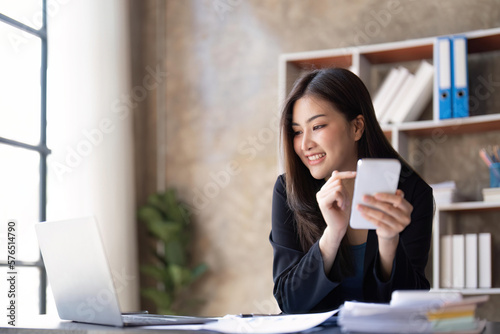 Asian businesswoman using smartphone to do accounting work with laptop and graph of financial reports in office