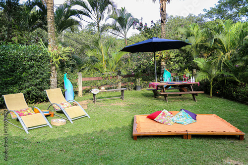 belo espaço ao ar livre em estilo zen para relaxamento, meditação e lazer com espreguiçadeiras, almofadas, mesa de piquenique de madeira e umbrelone em meio à vegetação tropical.