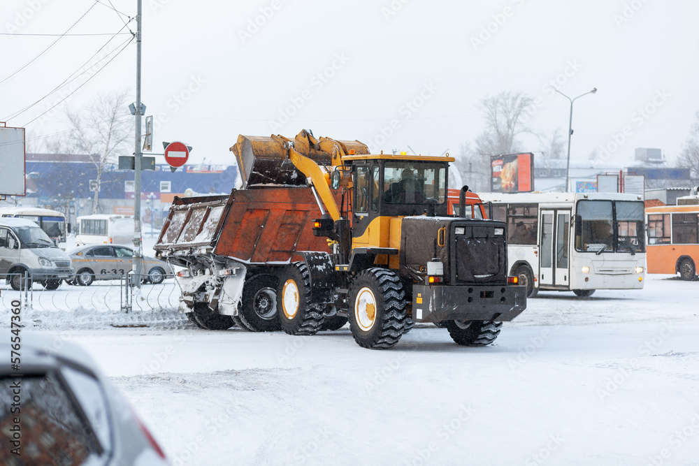 Orange tractor cleans up snow from the road and loads it into the truck. Cleaning and cleaning of roads in the city from snow in winter. Snow removal after snowfall and blizzards. Orange tractor cleans up snow from the road and loads it into the truck. Cleaning and cleaning of roads in the city from snow in winter. Snow removal after snowfall and blizzards.