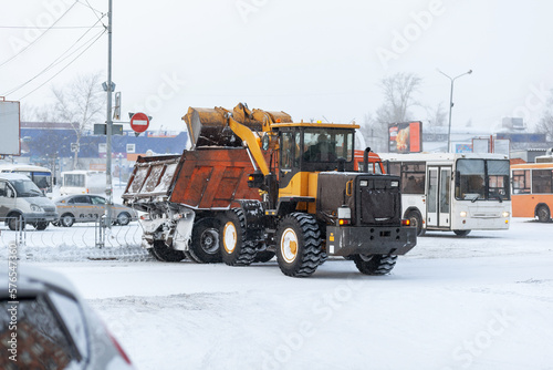 Orange tractor cleans up snow from the road and loads it into the truck. Cleaning and cleaning of roads in the city from snow in winter. Snow removal after snowfall and blizzards. 