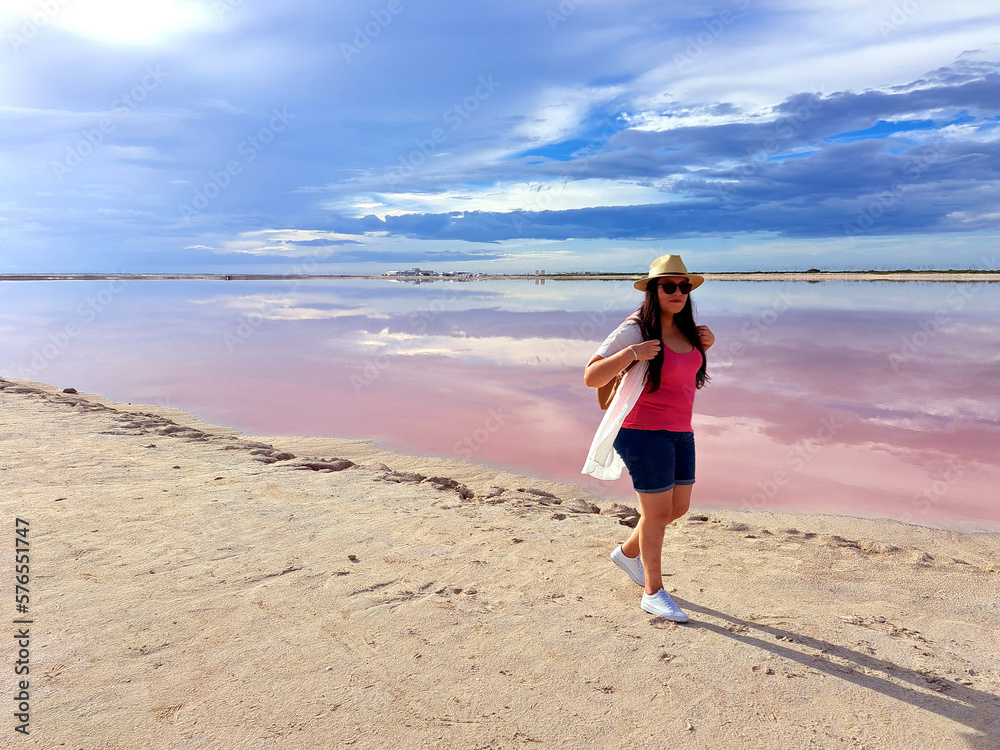 Latin adult woman with shorts, pink shirt, hat and sunglasses walks on the sand next to the pink colored lagoon with a high concentration of salt, Las Coloradas in Yucatan Mexico Latin adult woman with shorts, pink shirt, hat and sunglasses walks on the sand next to the pink colored lagoon with a high concentration of salt, Las Coloradas in Yucatan Mexico