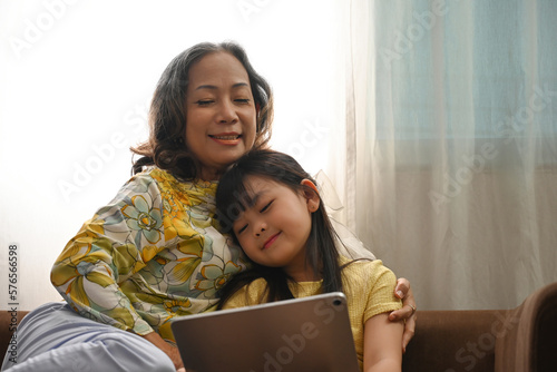 Happy senior grandma and cute little girl watching cartoons or playing game on digital tablet together.
