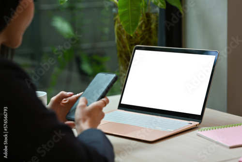 Side view of woman sitting front of laptop computer with blank screen and using smart phone.