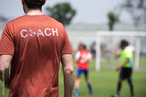 Male youth football coach standing and watching his team playing in a school tournament on a clear sky and sunny day. Sport, active lifestyle, happy activity and coaching concept. 