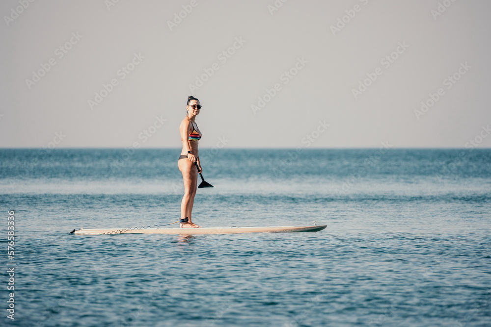 Sea woman sup. Silhouette of happy middle aged tanned woman in rainbow bikini, surfing on SUP board, confident paddling through water surface. Idyllic sunset. Active lifestyle at sea or river.
